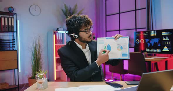 Businessman in Headset Sitting in front of Web Camera in Evening Office and Explaining report