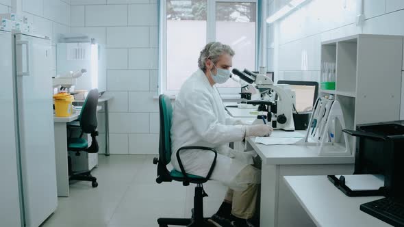 Laboratory Assistant Looks Through a Microscope and Writes Notes in a White Lab