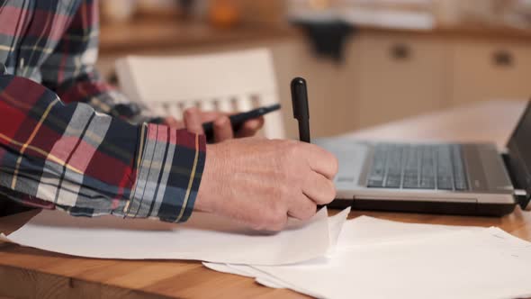 Elderly Man Working on Laptop and Writing Notes in Notebook Sitting at Table