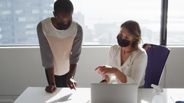 Two diverse female colleagues wearing face masks looking at laptop and discussing in office