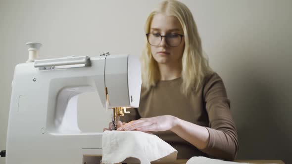 A Young Woman Sews Clothes Sitting in a Light Workshop at a Table with a Sewing Machine