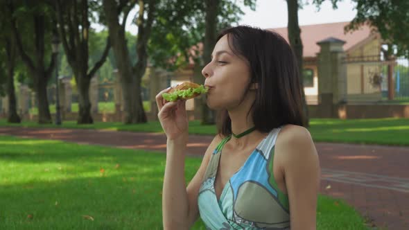 Girl Sitting on the Ground in the Park and Eating a Sandwich