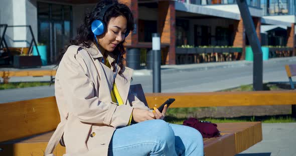 A Young Asian Woman in Headphones Listens to Music Through a Smartphone Application and Actively
