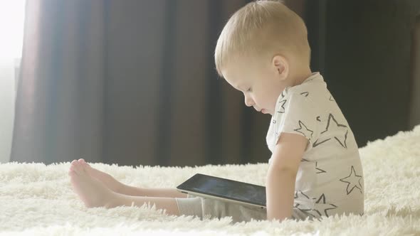 Little Boy Sitting on a Bed and Plays on the Tablet in the Room. Education and Technology Concept.