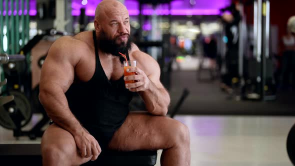 Adult Brawny Man with Large Biceps and Triceps is Resting in Gym Drinking Fresh Juice with Vitamin