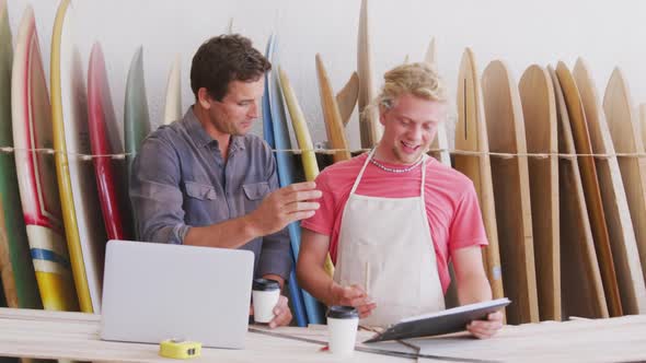 Two Caucasian male surfboard makers working on projects using a laptop computer