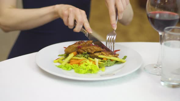 A Lady in an Evening Dress Eats Pork with Vegetables with a Fork and Knife.