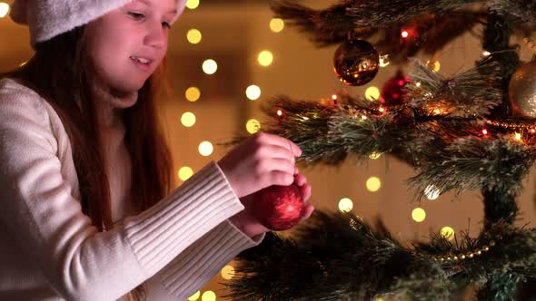Cute Little Girl in White Sweater and Santa Hat Hanging Decorative Toy Ball on Christmas Tree Branch alt