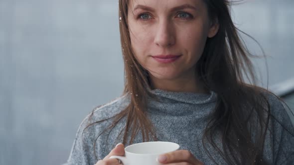 Caucasian Woman Stays on Balcony During Snowfall with Cup of Hot Coffee or Tea