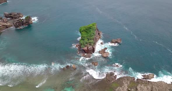 Drone shot of Coral island overgrown by green trees that are buffeted by the waves crashing in the t