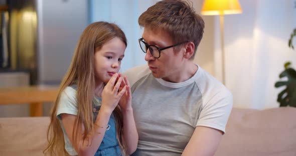 Joyful Dad Sitting with Little Girl on Couch Hugging and Talking