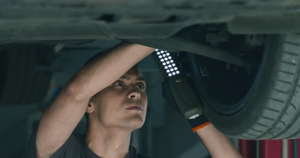 Young Auto Mechanic Holding Light and Examining Vehicle Suspension in Garage, Close Up
