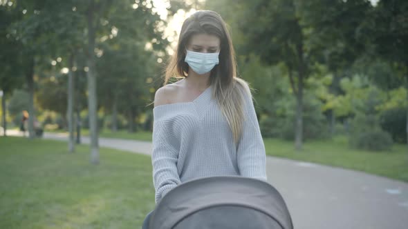 Confident Young Mother in Covid Face Mask Walking in Sunlight with Baby Stroller in Summer Park