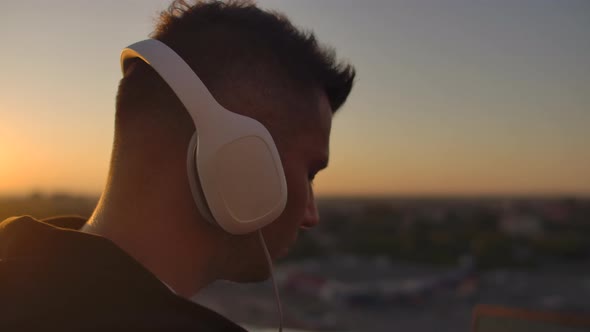 Close-up of a Man in Headphones Listening To Music on the Roof at Sunset with a View of the City.