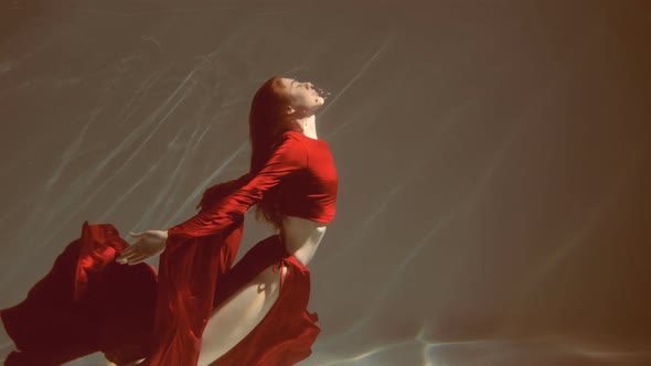 Attractive red-haired young woman swims beautifully underwater in a red dress. slow motion