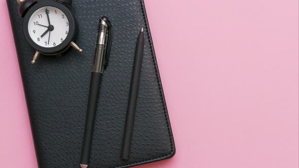 Black Notepad with a Pen and an Alarm Clock on a Pink Background