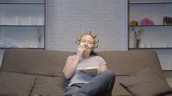 woman with facial mask eats cucumber while sitting on sofa 