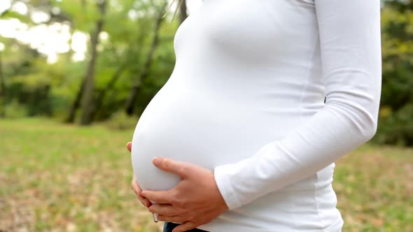 Young Pregnant Woman Stands Alone and Strokes Her Abdomen in Park - Closeup Belly