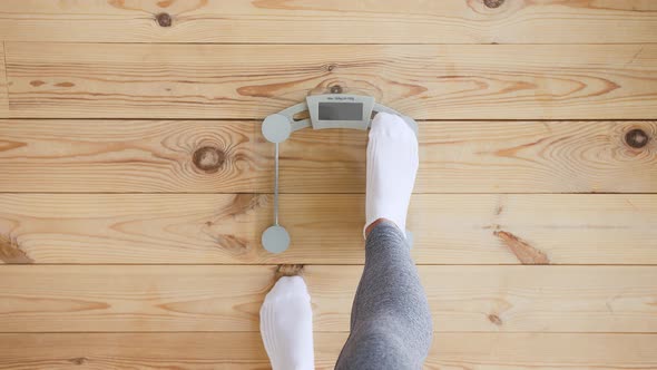 Unknown Female in Gray Leggings and White Socks is Standing on Weight Scale on Wooden Floor