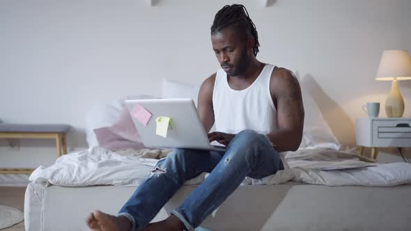 Wide Shot of Focused African American Freelancer Texting on Laptop Keyboard Sitting in Bedroom