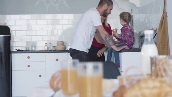 Cheerful Caucasian Parents Talking with Adorable Little Girl Sitting on Countertop in the Kitchen