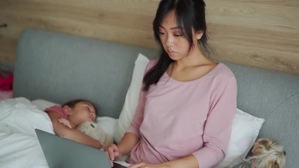 Serious Asian mom working on laptop while her daughter sleeping in the bed