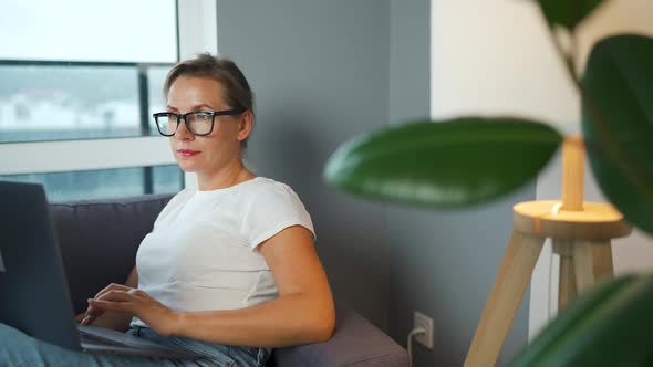 Woman with Glasses is Sitting on the Couch and Working on a Laptop
