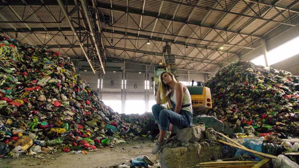 Young Woman in Jeans Sitting Against Operating Excavator Against Huge Pile of Trash