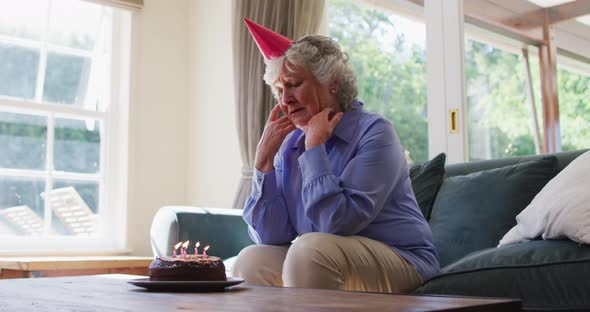Sad caucasian senior woman with birthday cake looking out of the window at home