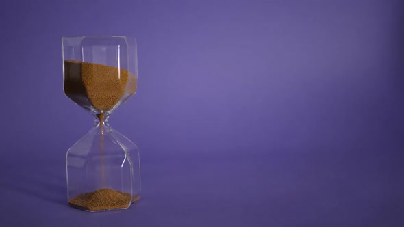 Glass Hourglass with Golden Sand on Purple Background