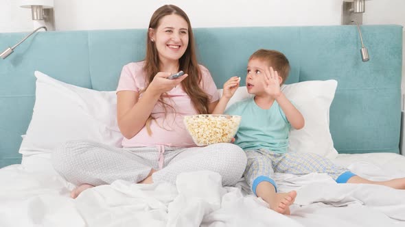 Happy Smiling Mother with Little Son Eating Popcorn and Watching Movie in Bed at Morning