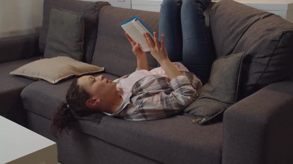 Adult Woman Reading Book Throwing Legs Over Back of Couch Indoors