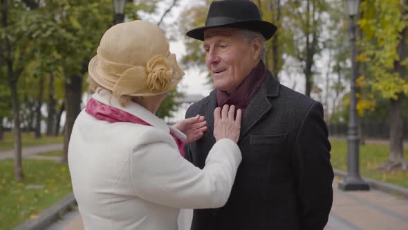 Mature Caucasian Woman in Elegant Cloche Hat and White Coat Correcting Clothes of Her Lovely Husband