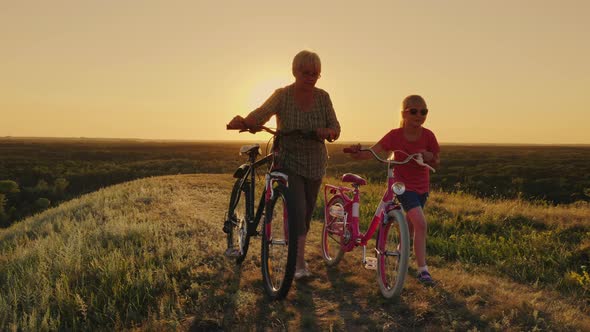 An Elderly Lady Walks with Her Granddaughter Bicycles at Sunset