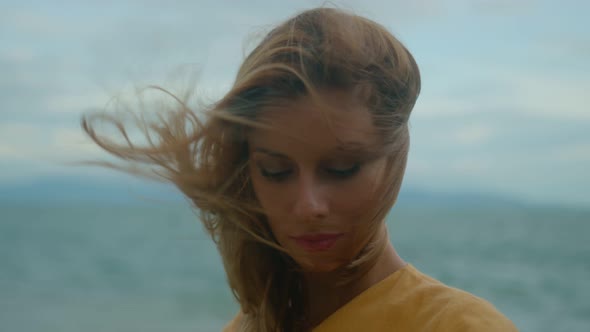 Beautiful Blonde in a Yellow Dress Alone on the Beach Looking at the Camera in Cloudy and Windy