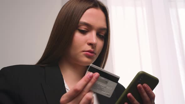 Portrait of young woman holding credit card and smartphone sitting on couch at home or office