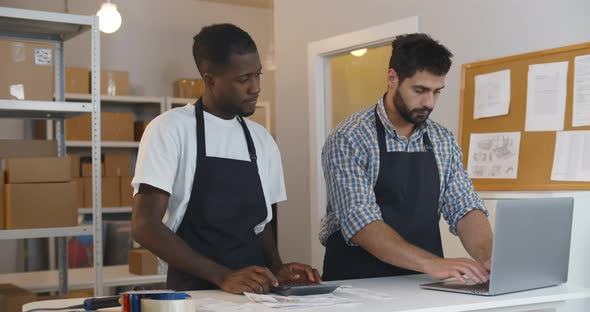 Small Business Owners Count Profit Using Laptop and Calculator in Warehouse