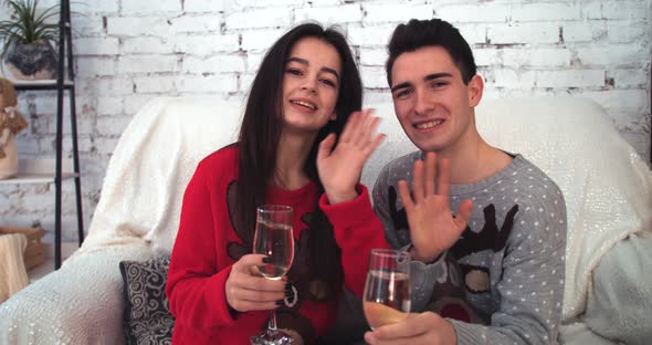 Attractive Girl with Boyfriend Waving at Camera Sitting on Sofa with Glasses in Hands