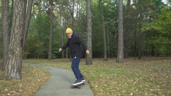 Adult Gray-haired Man Rides a Skateboard on a Path in a Forest Park, Autumn Time of the Year, Man