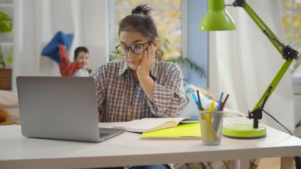 Young Tired Woman Trying To Work on Laptop at Home Office with Kids Playing on Background
