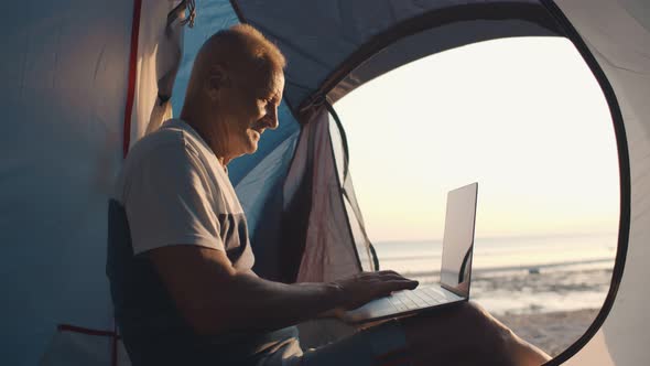 Side View of Senior Man Working on Laptop Sitting in Tent on Beach