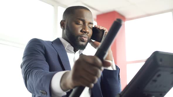 Tired Businessman Training on Stationary Bike and Talking on Smartphone at Gym