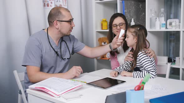 Friendly Doctor Talks with Patients Mom and Her Daughter. The Doctor Measures the Temperature of the alt
