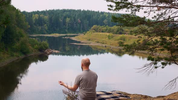 A Bald Male Meditating on the Edge of a Cliff Under a Tree Below is a Mountain River with a Calm
