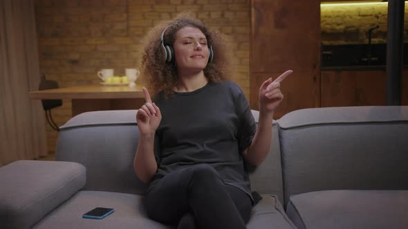 20s Young Woman Enjoys Listening Music in Wireless Headphones with Closed Eyes Sitting on Couch at