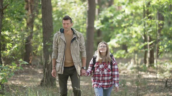 Happy Caucasian Man and Woman Holding Hands Walking with Backpacks in Sunny Forest Outdoors