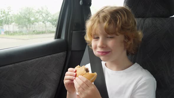 Blonde hungry boy eating a delicious cheeseburger in the car. alt
