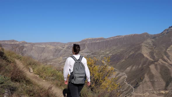 Young Woman in a White Shirt with a Backpack Climbs the Trail Against the Background of the