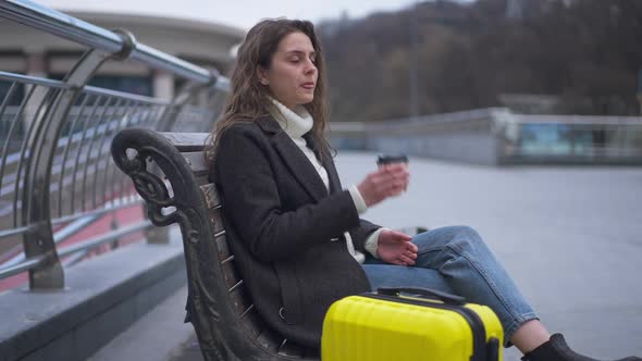 Side View Portrait of Gorgeous Confident Young Woman Sitting on Bench Drinking Coffee with Luggage