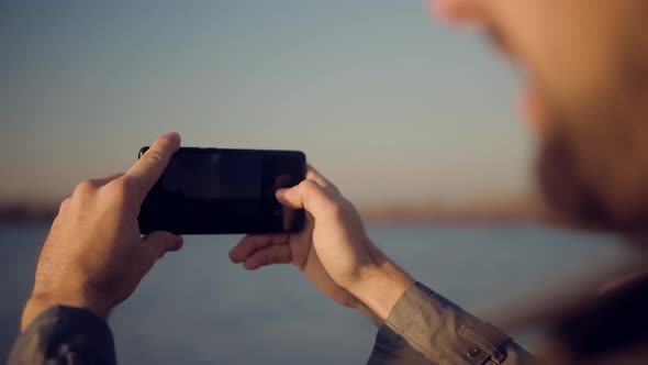 Man Hands Holding Mobile Phone And Taking Picture At Sunset. Guy Taking Photo of Sunset With Mobile